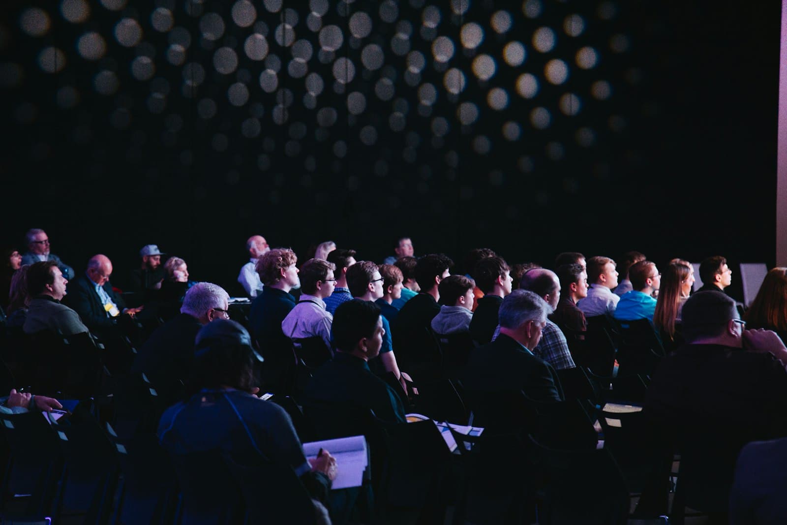 An audience seated in rows facing a speaker on a lit stage